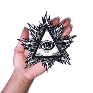 Puede incluir: Parche bordado en blanco y negro de un ojo que todo lo ve dentro de un triángulo con llamas alrededor.