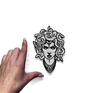 Könnte beinhalten: Schwarz-weiß gestickter Aufnäher von Medusa, einer mythischen griechischen Figur mit Schlangenhaaren, die eine Halskette trägt.