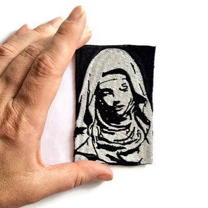 May include: Black and white embroidered patch of a woman wearing a hooded robe. The patch is on a black background.