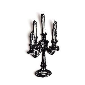 Può includere: Toppa ricamata in bianco e nero di un candelabro con tre candele accese. Il candelabro ha un design dettagliato con una base rotonda e bracci curvi che sostengono le candele. Le candele presentano dettagli di cera fusa.