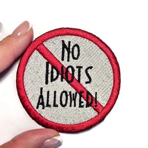 May include: A round embroidered patch with a red border and a gray center. The text "NO IDIOTS ALLOWED!" is in black, with a red diagonal line through the center.