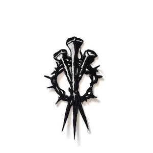 May include: Black embroidered patch featuring three nails and a thorny wreath design. The patch is a detailed, gothic-inspired emblem, suitable for jackets, bags, or other fabric items. The design is set against a white background.