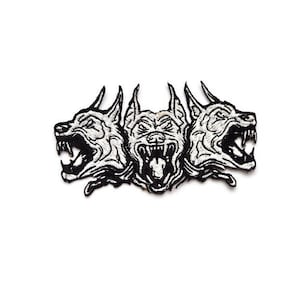 Puede incluir: Parche bordado en blanco y negro que representa un perro de tres cabezas con la boca abierta. El parche tiene costuras detalladas, que muestran los dientes y el pelaje del perro. El diseño está sobre un fondo blanco.