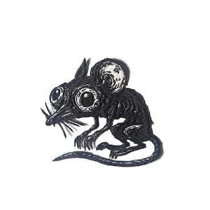 Puede incluir: Parche bordado negro de un ratón estilizado con ojos grandes y una larga cola. El ratón tiene costuras detalladas, lo que le da una apariencia texturizada. El diseño está sobre un fondo blanco.