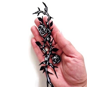K&ouml;nnte beinhalten: Schwarz-wei&szlig;er, gestickter Aufn&auml;her mit einem Rosenranken-Design. Der Aufn&auml;her hat zwei Rosen, Bl&auml;tter und Dornen. Das Design wird in einer Hand vor wei&szlig;em Hintergrund gehalten.