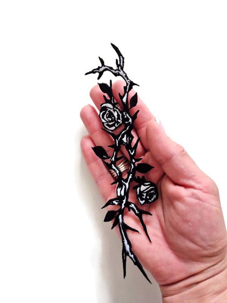 K&ouml;nnte beinhalten: Schwarz-wei&szlig;er, gestickter Aufn&auml;her mit einem Rosenranken-Design. Der Aufn&auml;her hat zwei Rosen, Bl&auml;tter und Dornen. Das Design wird in einer Hand vor wei&szlig;em Hintergrund gehalten.