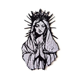 Può includere: Toppa ricamata in bianco e nero raffigurante una figura incoronata con le mani giunte in preghiera. Il disegno comprende un'aureola e tratti del viso dettagliati, su sfondo bianco. La toppa ha un aspetto strutturato.