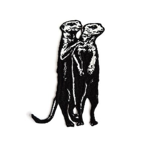 Puede incluir: Parche bordado en blanco y negro que representa dos suricatos de pie y juntos. El parche tiene un aspecto texturizado debido a las costuras. Los suricatos se destacan sobre un fondo blanco liso.