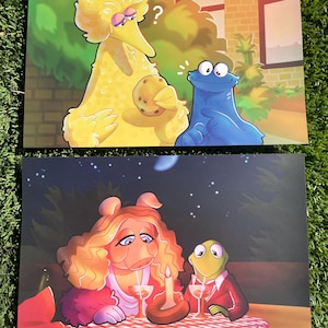 May include: Two art prints featuring cartoon characters. The top print shows Big Bird holding a cookie, with Cookie Monster looking on. The bottom print depicts Miss Piggy and Kermit the Frog at a candlelit dinner. The prints are on a green surface.