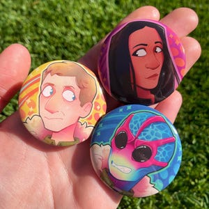 May include: Three colourful round badges with cartoon portraits. One badge features a man with light brown hair and blue eyes, another depicts a woman with dark hair, and the third shows a character with pink glasses.