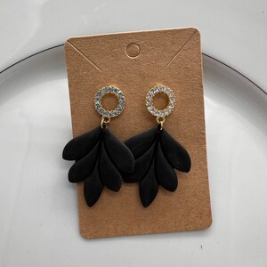 May include: Black dangle earrings with a gold circle adorned with clear rhinestones and black leaf-shaped accents.