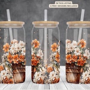May include: Three clear glass tumblers with lids and straws. Each tumbler features a floral design with orange, white, and green flowers in a brown wooden bucket. The background is a blurred image of a gray wall.