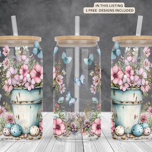 Puede incluir: Tres vasos de vidrio transparente con una pajita y un diseño floral. El diseño presenta flores rosas y blancas en un cubo azul y blanco con mariposas y hojas verdes. Los vasos están sobre una superficie blanca.