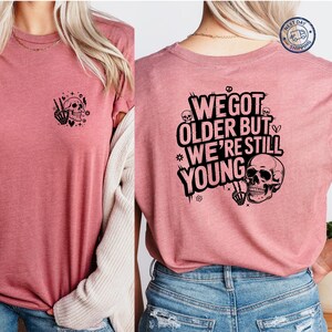 May include: A dusty rose t-shirt with a black skull graphic on the front and back. The front graphic features a skull with a peace sign hand gesture. The back of the shirt says "We Got Older But We're Still Young."