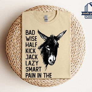 May include: A tan t-shirt with a black and white illustration of a donkey's head. The text "Bad Wise Half Kick Jack Lazy Smart Pain in the" is printed on the shirt.