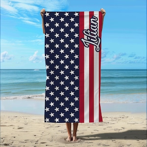 May include: A beach towel featuring the American flag design with a blue field of white stars and red and white stripes. The name "Lilian" is written in a cursive font on the right side of the towel. The towel is held up on a beach with the ocean and sky in the background.