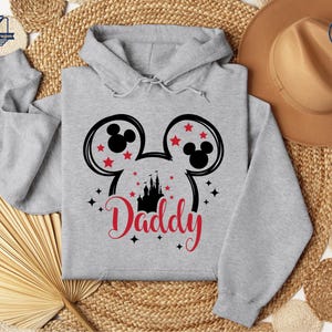 Puede incluir: Una sudadera con capucha gris claro con un dise&ntilde;o de cabeza de Mickey Mouse negro y estrellas rojas. La palabra "Daddy" est&aacute; escrita en rojo debajo de una silueta de castillo negro. La sudadera tiene una capucha con cord&oacute;n.