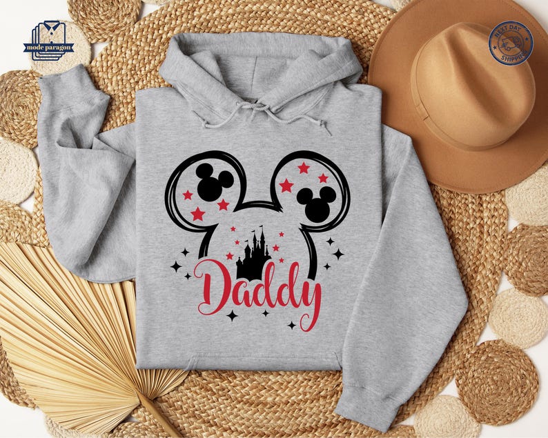 Puede incluir: Una sudadera con capucha gris claro con un dise&ntilde;o de cabeza de Mickey Mouse negro y estrellas rojas. La palabra "Daddy" est&aacute; escrita en rojo debajo de una silueta de castillo negro. La sudadera tiene una capucha con cord&oacute;n.