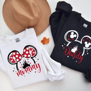 Puede incluir: Dos sudaderas, una blanca y otra negra, con dise&ntilde;os tem&aacute;ticos de Disney. La sudadera blanca presenta un lazo de Minnie Mouse con corazones y lunares rojos, la palabra "mommy" en rojo y una silueta de castillo. La sudadera negra tiene un dise&ntilde;o de cabeza de Mickey Mouse con la palabra "Daddy".