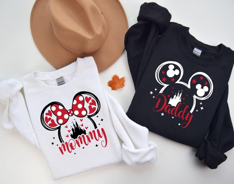 Puede incluir: Dos sudaderas, una blanca y otra negra, con dise&ntilde;os tem&aacute;ticos de Disney. La sudadera blanca presenta un lazo de Minnie Mouse con corazones y lunares rojos, la palabra "mommy" en rojo y una silueta de castillo. La sudadera negra tiene un dise&ntilde;o de cabeza de Mickey Mouse con la palabra "Daddy".