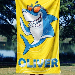 May include: Yellow beach towel featuring a cartoon shark wearing sunglasses and waving. The shark is blue and white with a big smile. The name "OLIVER" is printed in blue on the bottom of the towel.