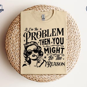 May include: A beige t-shirt with a black and white graphic print. The graphic features a woman with sunglasses and a drink, and the text "If I'm the PROBLEM THEN YOU MIGHT Be The REASON".
