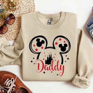 Puede incluir: Una sudadera beige con un dise&ntilde;o de Mickey Mouse en negro y rojo y la palabra "Daddy" en escritura roja. El dise&ntilde;o incluye orejas de Mickey Mouse, estrellas rojas y una silueta de castillo. La sudadera es de estilo casual.