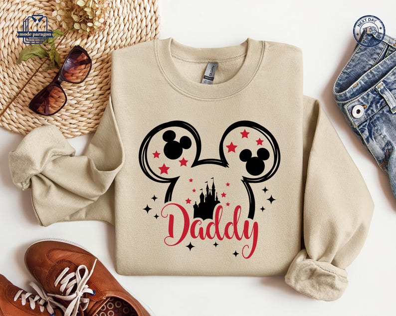 Puede incluir: Una sudadera beige con un dise&ntilde;o de Mickey Mouse en negro y rojo y la palabra "Daddy" en escritura roja. El dise&ntilde;o incluye orejas de Mickey Mouse, estrellas rojas y una silueta de castillo. La sudadera es de estilo casual.