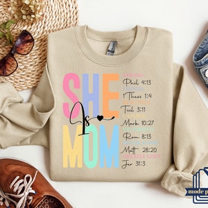 May include: A beige sweatshirt with the text "SHE is MOM" in rainbow colors. The text is surrounded by bible verses in black text.
