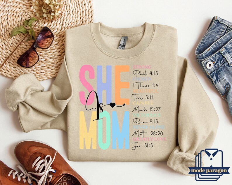May include: A beige sweatshirt with the text "SHE is MOM" in rainbow colors. The text is surrounded by bible verses in black text.