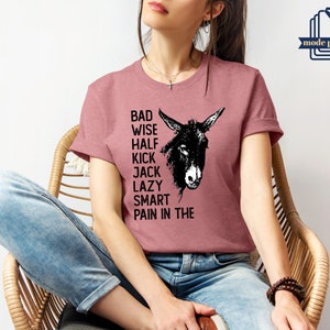 May include: A woman wearing a dusty rose t-shirt with a black donkey graphic and the text "Bad Wise Half Kick Jack Lazy Smart Pain in the".
