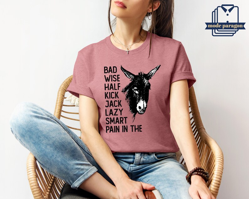 May include: A woman wearing a dusty rose t-shirt with a black donkey graphic and the text "Bad Wise Half Kick Jack Lazy Smart Pain in the".
