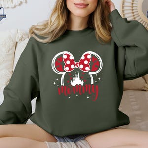 Puede incluir: Sudadera verde oscuro con un dise&ntilde;o de Minnie Mouse. Presenta un lazo rojo con lunares, orejas rojas en forma de coraz&oacute;n y la palabra "mam&aacute;" en escritura roja. Una silueta de castillo blanco est&aacute; debajo de las orejas. La sudadera es casual.
