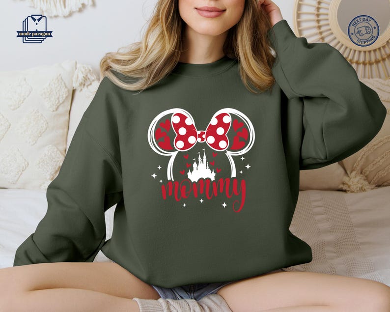 Puede incluir: Sudadera verde oscuro con un dise&ntilde;o de Minnie Mouse. Presenta un lazo rojo con lunares, orejas rojas en forma de coraz&oacute;n y la palabra "mam&aacute;" en escritura roja. Una silueta de castillo blanco est&aacute; debajo de las orejas. La sudadera es casual.