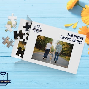 May include: A white puzzle box with the text "300 Pieces Custom Design" and a family photo. Several puzzle pieces are scattered around the box. Yellow flowers are in the background. The box has the "mode paragon" logo.