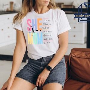 May include: White t-shirt with the text "SHE is MOM" in rainbow colors. The text is accompanied by bible verses, including "STRONG Phil 4:13", "HOPE 1 Thess 1:4", "BEAUTIFUL Tit 3:11", "GRACE Mark 10:27", "JOY Rom 8:13", "LOVE Matt 28:20", "GREATLY LOVE Jer 31:3".