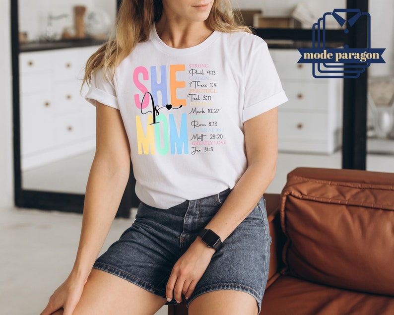 May include: White t-shirt with the text "SHE is MOM" in rainbow colors. The text is accompanied by bible verses, including "STRONG Phil 4:13", "HOPE 1 Thess 1:4", "BEAUTIFUL Tit 3:11", "GRACE Mark 10:27", "JOY Rom 8:13", "LOVE Matt 28:20", "GREATLY LOVE Jer 31:3".