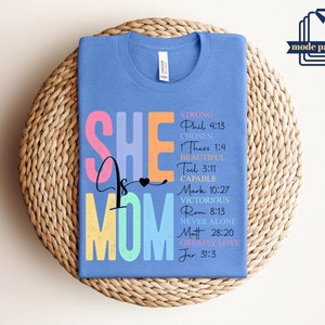 May include: A light blue t-shirt with the text "She is Mom" in rainbow colors. The text is surrounded by bible verses about strength, love, and being chosen.