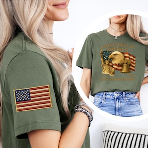 Puede incluir: Camiseta verde oliva con un parche de la bandera estadounidense en la manga. La camiseta presenta una impresi&oacute;n gr&aacute;fica con el texto "United States of America" e im&aacute;genes de la Estatua de la Libertad, un &aacute;guila, una campana y la bandera estadounidense.