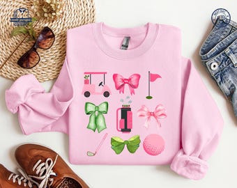 May include: Pink sweatshirt with a golf-themed design featuring a golf cart, golf clubs, a golf ball, and bows. The design is in shades of pink, green, and black.