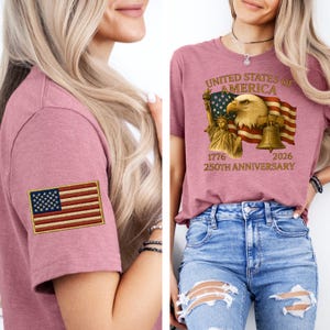 Puede incluir: Dos camisetas de color malva. Una presenta una bandera estadounidense bordada en la manga. La otra muestra un gr&aacute;fico con un &aacute;guila, la Estatua de la Libertad, una campana y el texto "UNITED STATES OF AMERICA 1776 2026 250TH ANNIVERSARY".