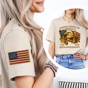 Puede incluir: Camiseta beige con un parche bordado de la bandera estadounidense en la manga izquierda. La camiseta presenta un gr&aacute;fico en la parte delantera con el texto "United States of America" y las fechas "1776" y "2026" conmemorando el 250 aniversario.