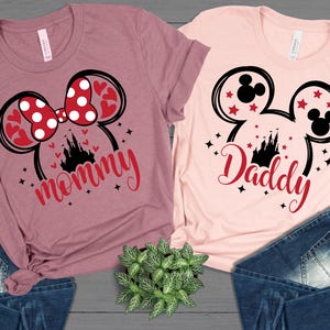 Puede incluir: Dos camisetas con dise&ntilde;os tem&aacute;ticos de Disney. Una es malva con un lazo de Minnie Mouse y la palabra "mam&aacute;" en escritura roja. La otra es rosa claro con orejas de Mickey Mouse y la palabra "pap&aacute;" en escritura roja. Ambas camisetas tienen un castillo.