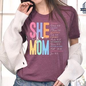 May include: A maroon t-shirt with the text "SHE is MOM" in rainbow colors. The text is surrounded by a list of words and bible verses describing the qualities of a mother.
