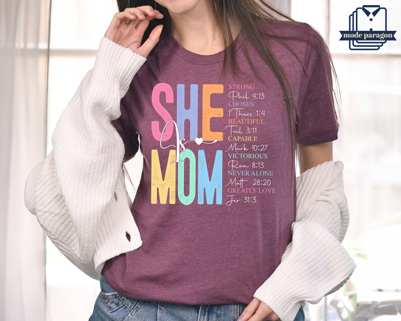 May include: A maroon t-shirt with the text "SHE is MOM" in rainbow colors. The text is surrounded by a list of words and bible verses describing the qualities of a mother.