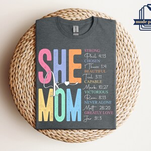 May include: A dark gray t-shirt with the text "SHE IS MOM" in colorful letters. The text is surrounded by bible verses, including "Strong Phil 4:13", "Chosen 1 Thess 1:4", "Beautiful Teel 3:11", "Capable Mark 10:27", "Victorious Rom 8:13", "Never Alone Matt 28:20", and "Greatly Love Jer 31:3".
