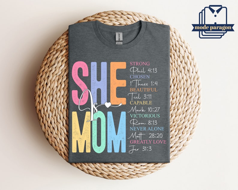 May include: A dark gray t-shirt with the text "SHE IS MOM" in colorful letters. The text is surrounded by bible verses, including "Strong Phil 4:13", "Chosen 1 Thess 1:4", "Beautiful Teel 3:11", "Capable Mark 10:27", "Victorious Rom 8:13", "Never Alone Matt 28:20", and "Greatly Love Jer 31:3".