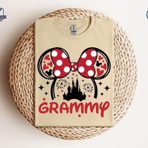 May include: A tan t-shirt with a black silhouette of Cinderella's castle and a red and white Minnie Mouse bow. The text "GRAMMY" is printed in red below the image.