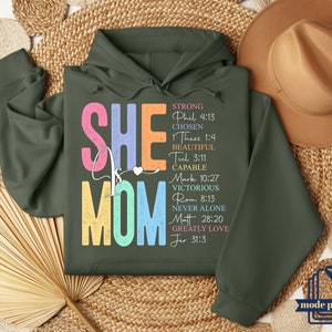 May include: A green hooded sweatshirt with the words "She is Mom" in colorful letters. The words "Strong, Chosen, Beautiful, Capable, Victorious, Never Alone, Greatly Love" are listed vertically with corresponding Bible verses.