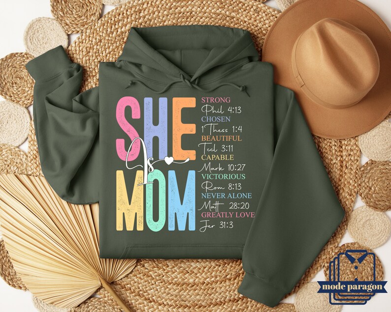 May include: A green hooded sweatshirt with the words "She is Mom" in colorful letters. The words "Strong, Chosen, Beautiful, Capable, Victorious, Never Alone, Greatly Love" are listed vertically with corresponding Bible verses.